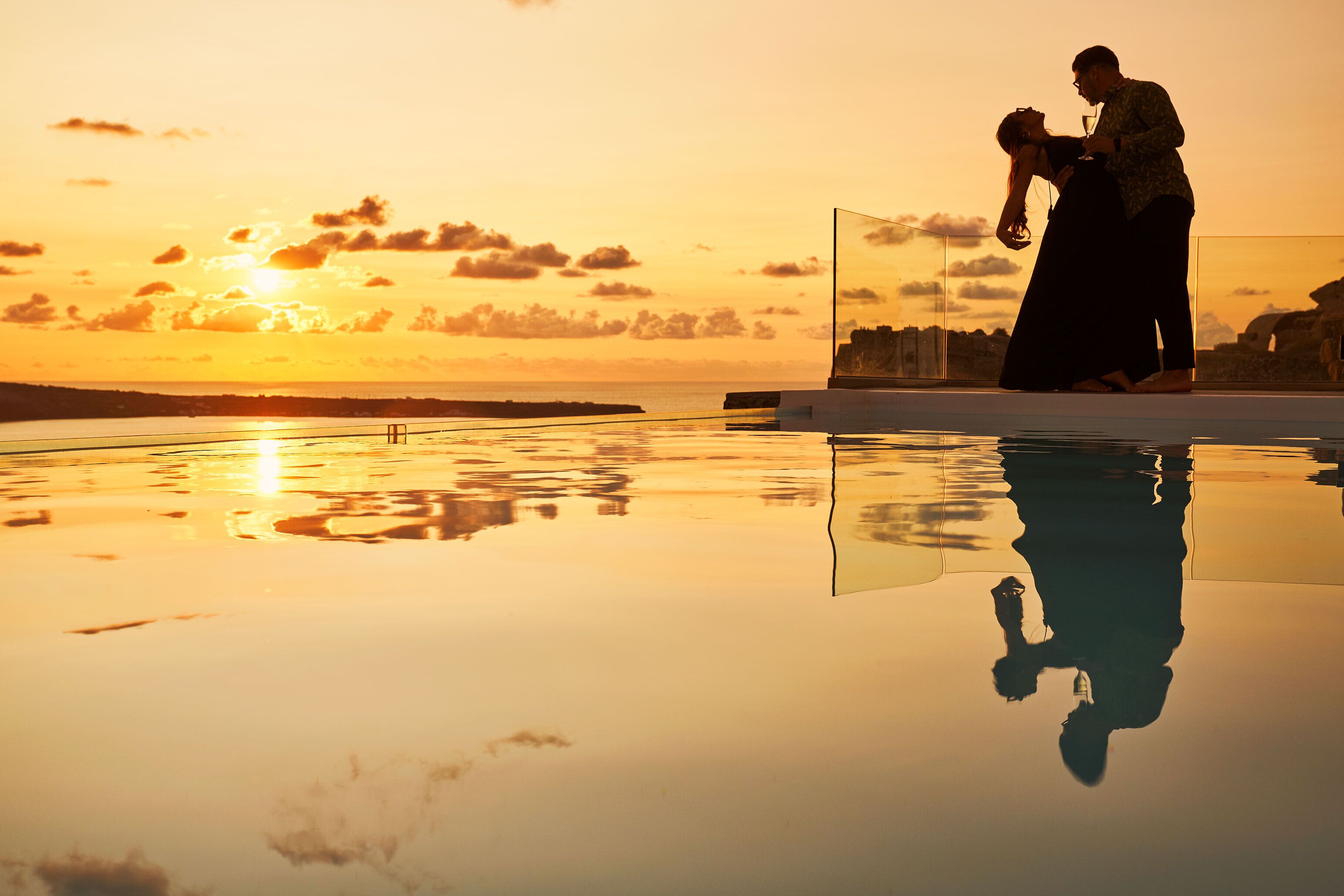 Una pareja parada junto a un estanque reflectante al atardecer, con un paisaje urbano de fondo.