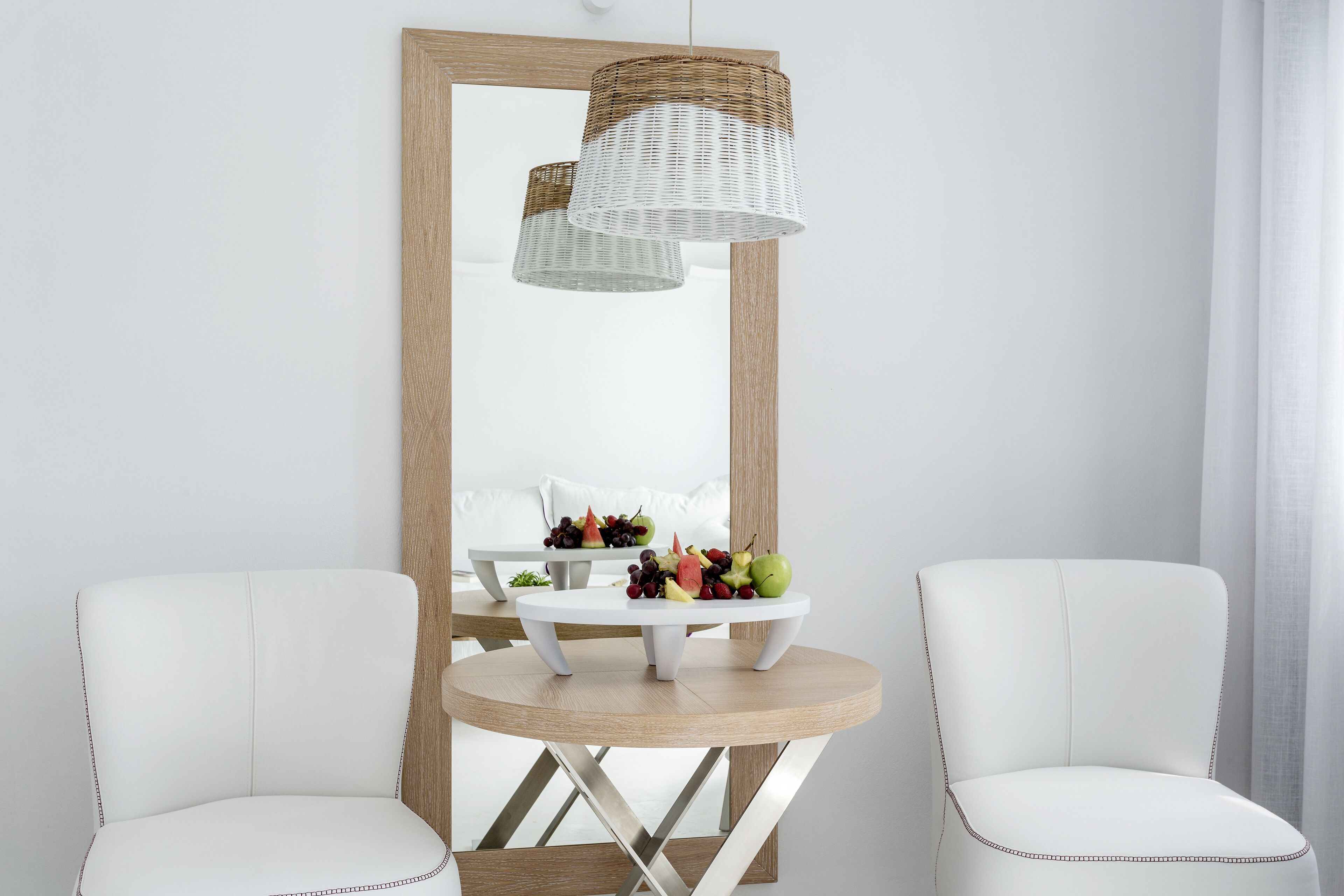 A modern room with a round wooden table, two white leather chairs, a wicker pendant light, and a mirror reflecting the room.