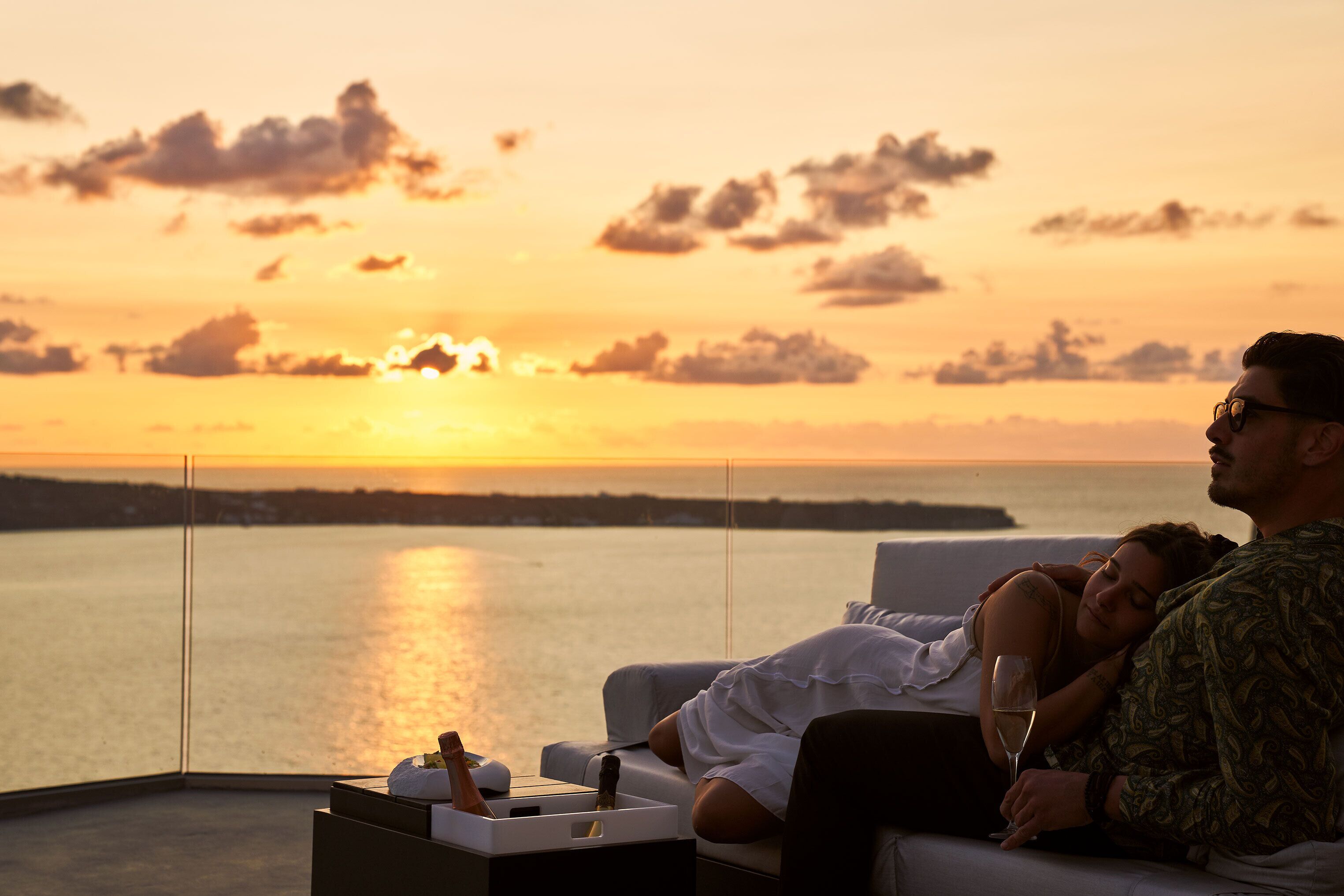 Um casal relaxando em uma varanda com vista para o pôr do sol sobre a água.
