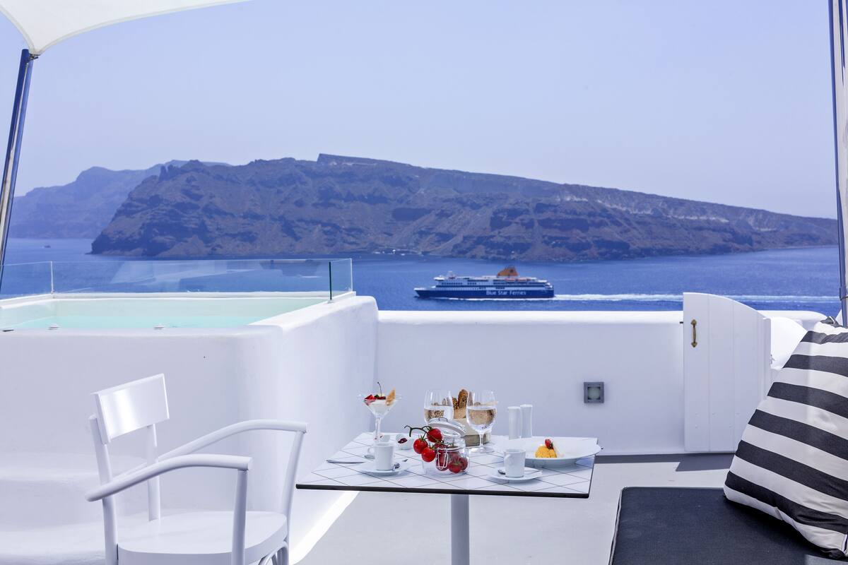 A sunlit terrace with a table set for dining, overlooking a calm sea and a rocky island.