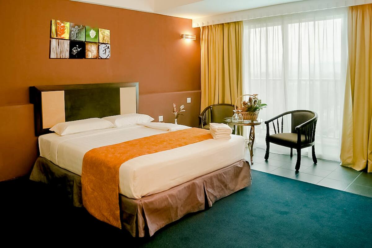 A hotel room with a large bed, a chair, a small table with a plant, and a window with curtains.