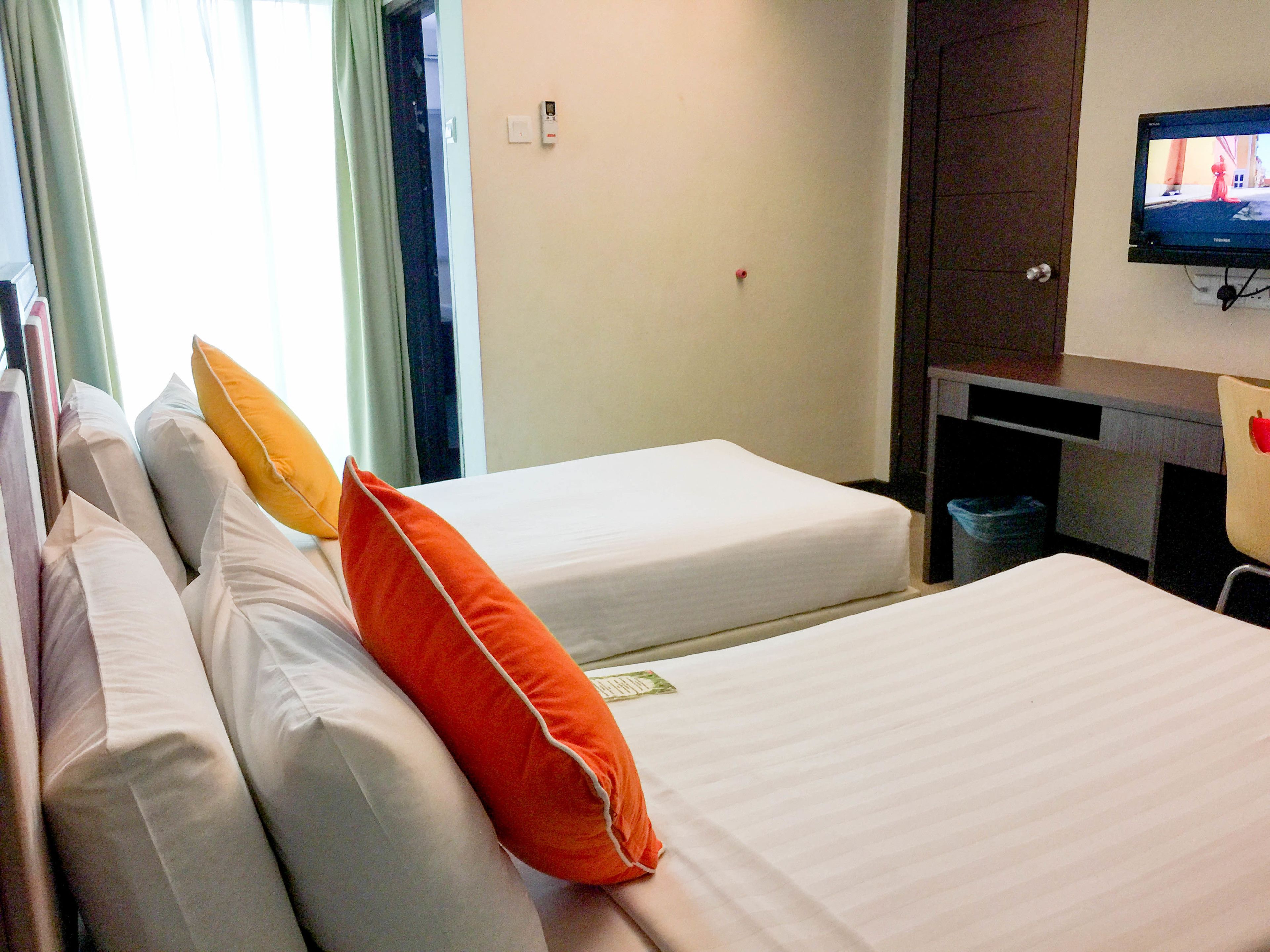 A hotel room with two beds, each with white bedding and orange pillows. There is a television mounted on the wall, a desk with a chair, and a trash bin.