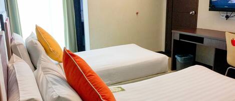 A hotel room with two beds, each with white bedding and orange pillows. There is a television mounted on the wall, a desk with a chair, and a trash bin.