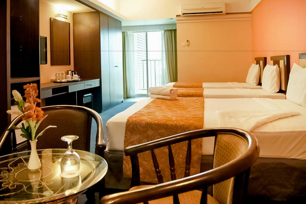 A hotel room with a desk, two beds, a chair, a table with a vase, and a window with curtains.