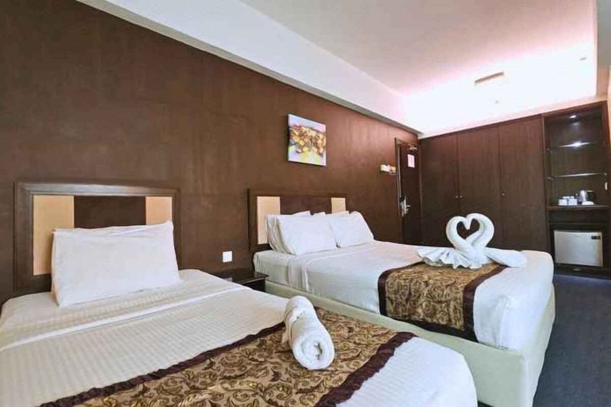 Habitación de hotel con dos camas, cada una con cabecera, mesita de noche y lámpara. Cuenta con un armario y una pequeña zona de estar con una silla y una mesa.