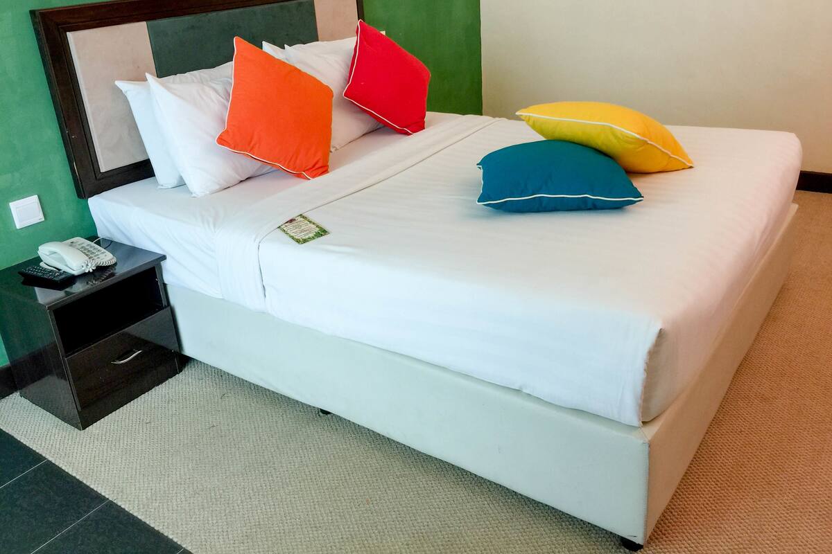 A neatly made bed with white bedding and colorful pillows, a bedside table with a phone and remote control, and a green wall in the background.