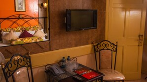 A hotel room with a bed with patterned pillows, a bedside table with a phone and a red booklet, a TV mounted on the wall, and a door. - Kahama Hotel (Nairobi)