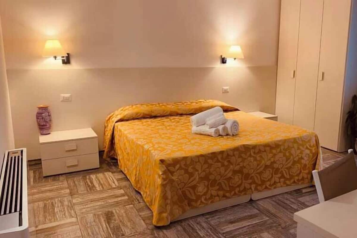 Una habitación de hotel con cama, mesitas de noche, una planta en maceta y un escritorio.