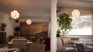 A restaurant interior with wooden tables and chairs, hanging paper lamps, a large painting of a sunset, and potted plants. - Hotel Mûita di Mari (Santa Teresa di Gallura)
