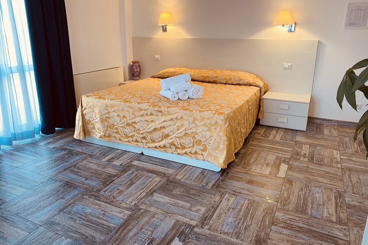Una habitación de hotel con una cama, dos lámparas de noche, una mesita de noche, un jarrón y una planta en maceta.
