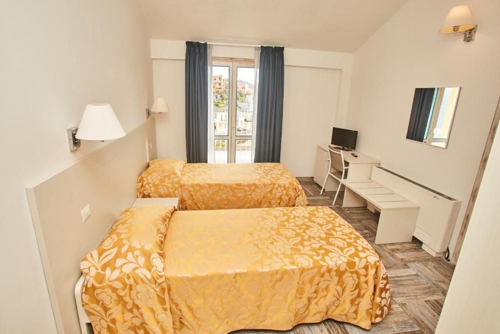 A hotel room with two beds, a desk, a TV, and a window with curtains.