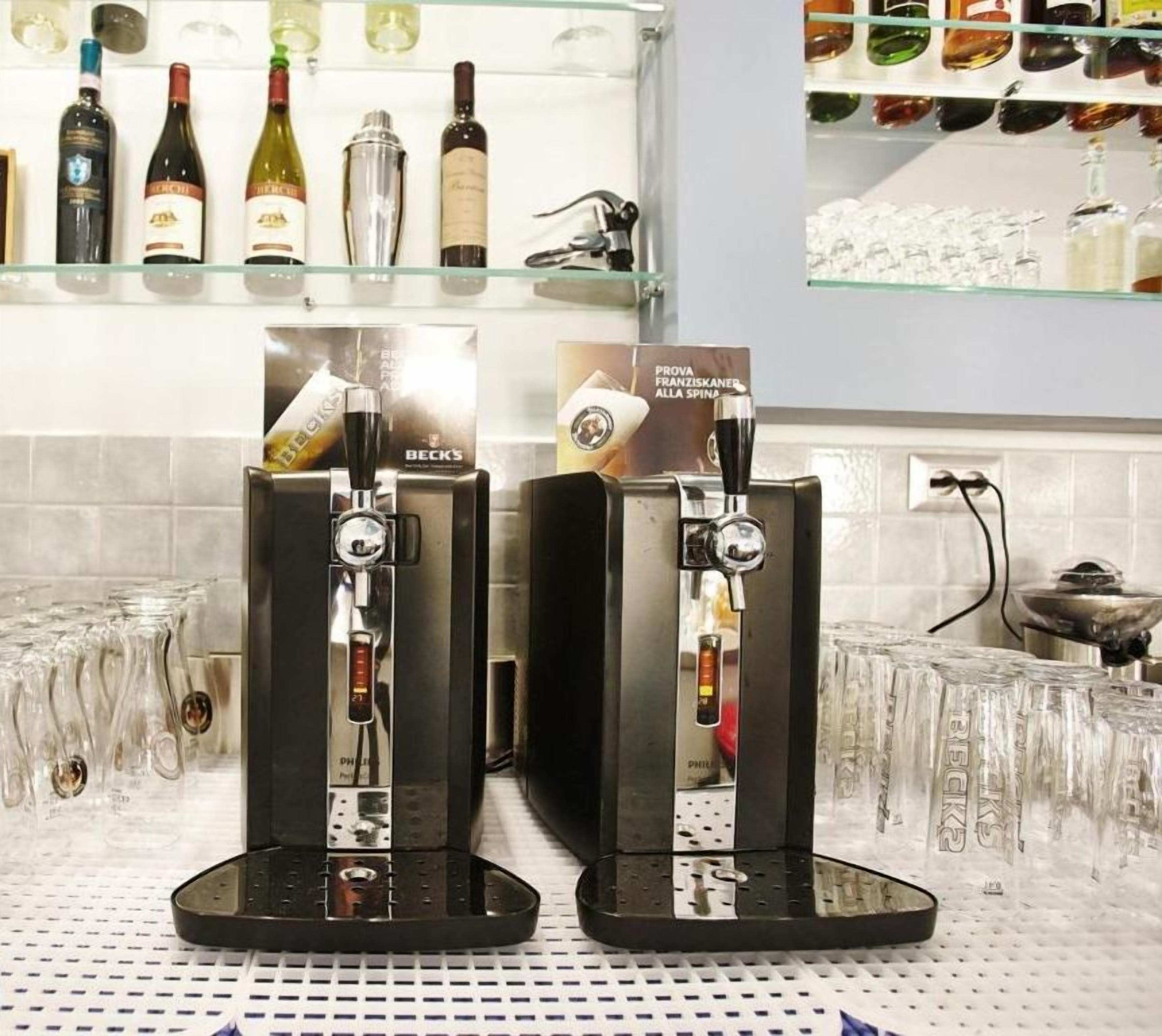 Dos máquinas de café con bandejas recuperadoras, un estante de vidrio con botellas de vino, y el logotipo de Hotel Mulita di Mori.