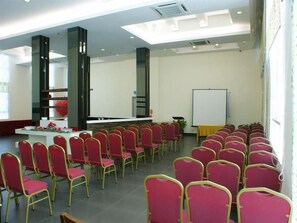 Un salón de conferencias con filas de sillas rojas, una pizarra y una larga mesa con arreglos florales.