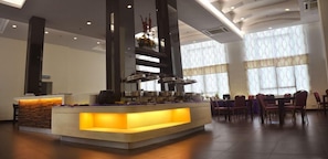 Un'area reception di un hotel con un banco centrale, diverse postazioni per il buffet e una sala da pranzo con tavoli e sedie.