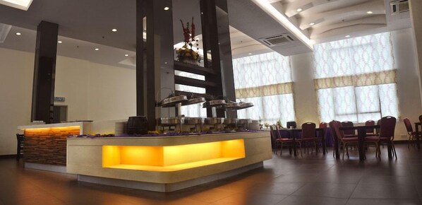 Zona de recepción de hotel con un mostrador central, varias estaciones de buffet y un comedor con mesas y sillas.