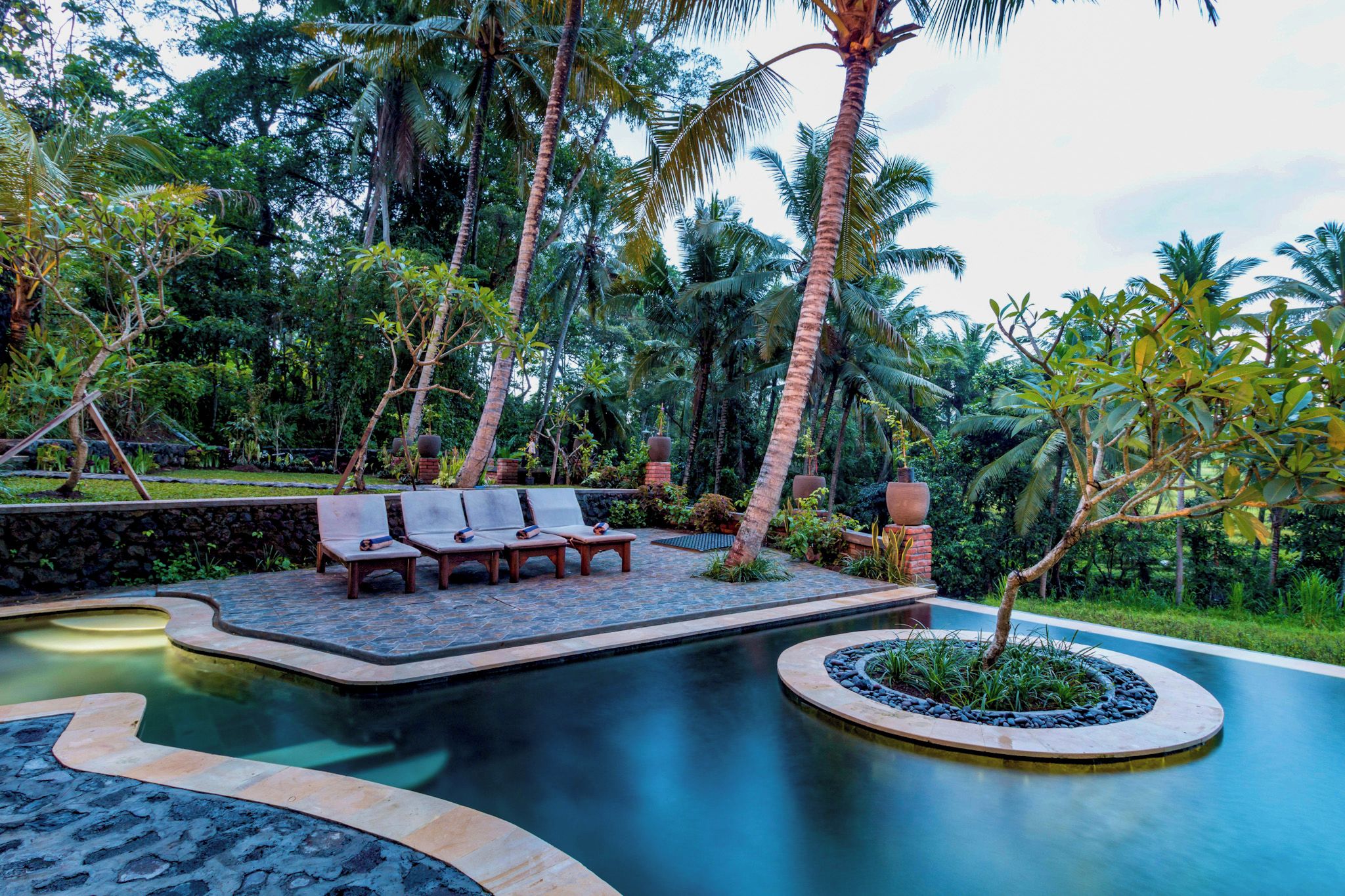 Un área de piscina con sillones de descanso y una fuente circular rodeada de vegetación tropical.