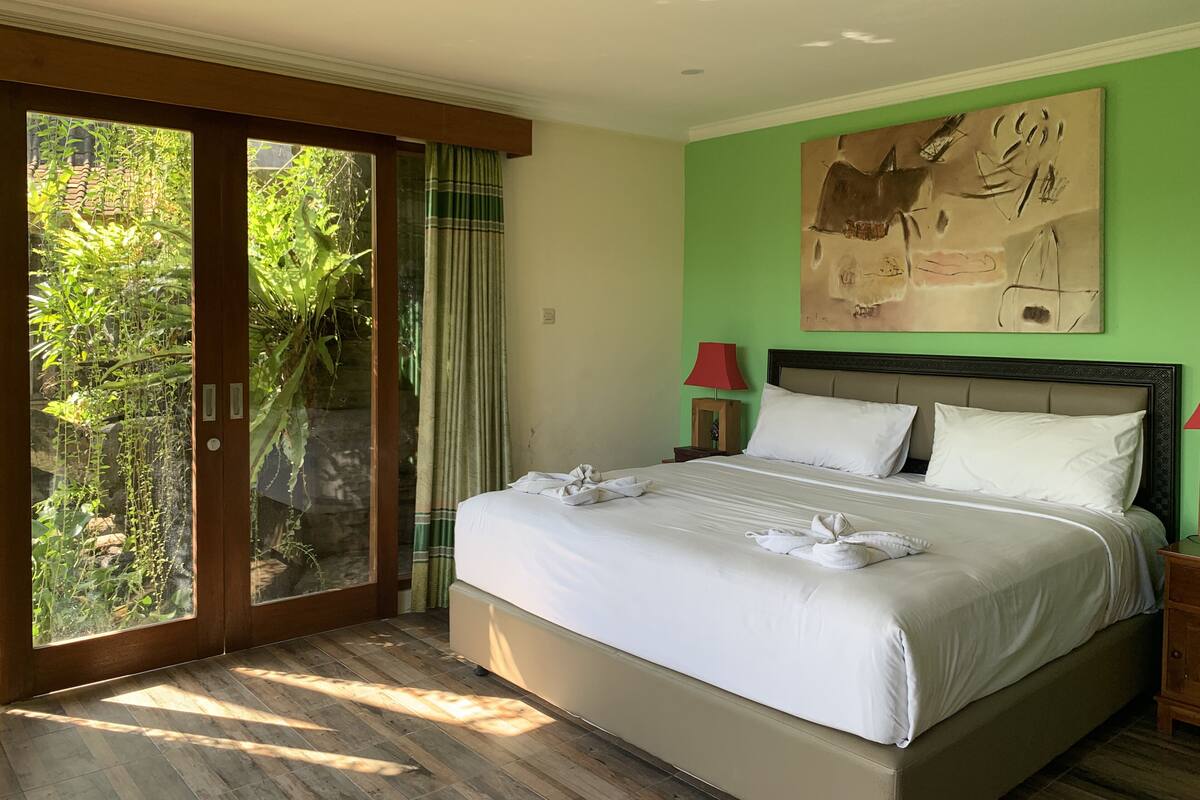 A bedroom with a large bed, a sliding glass door leading to a garden, a red lamp, and a painting on the wall.