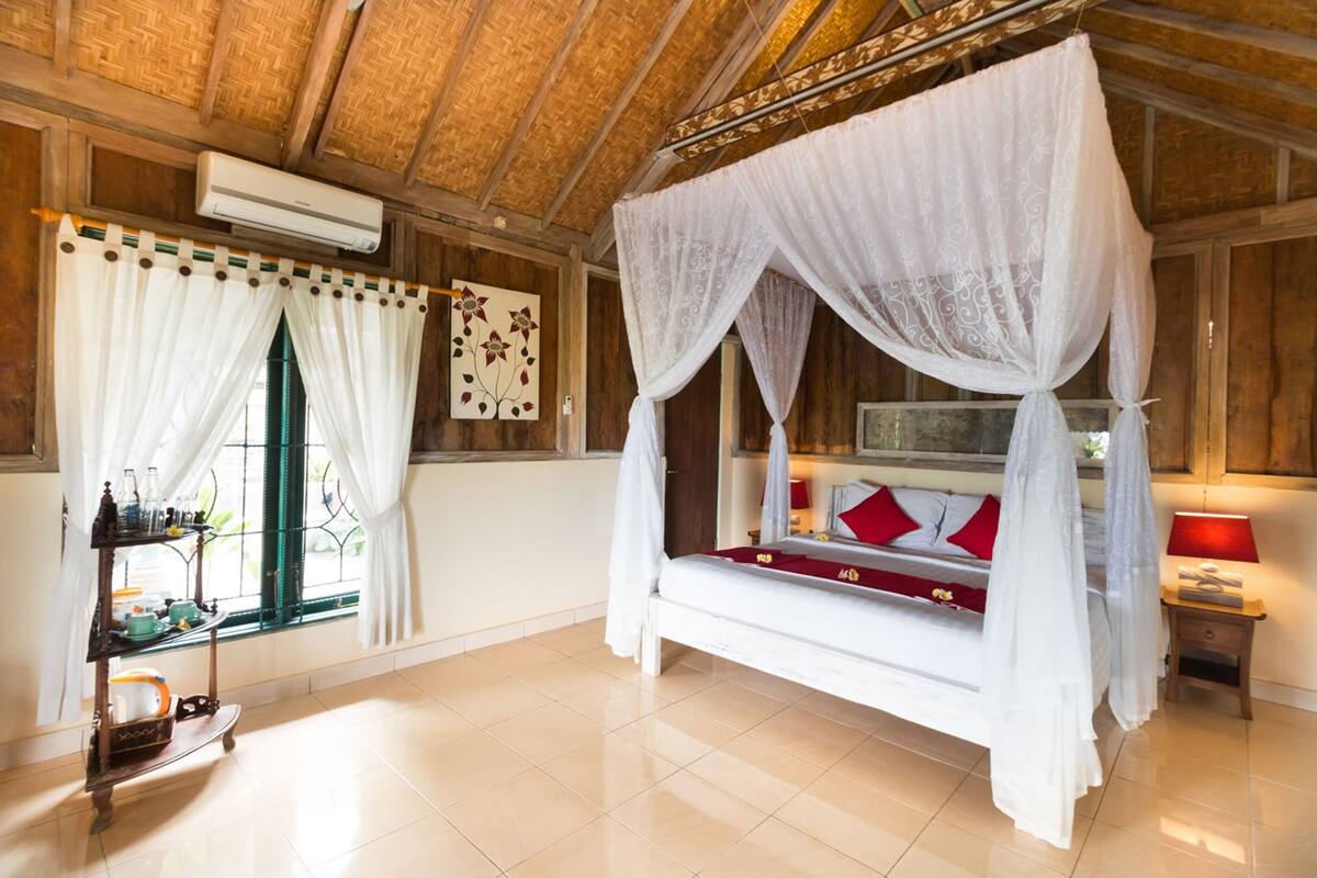 A bedroom with a four-poster bed, a window with curtains, a bedside table with a lamp, and a wall-mounted air conditioner.