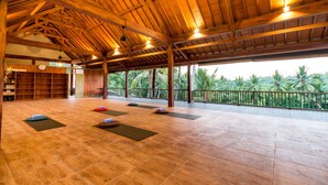 A spacious indoor area with a high ceiling, wooden beams, and large windows offering a view of palm trees. - Umasari Rice Terrace Villa (Mengwi)