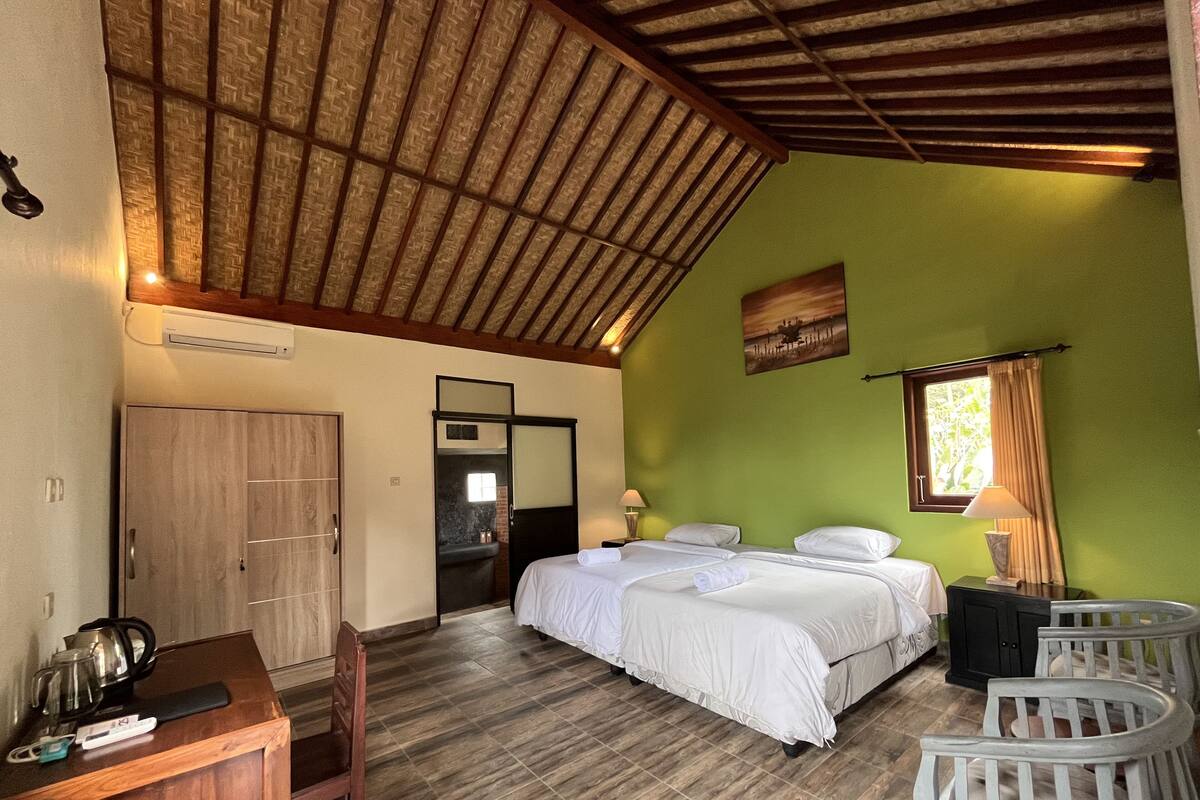 A bedroom with a bed, a desk, and a chair. There is a window with curtains, a painting on the wall, and a wooden ceiling.