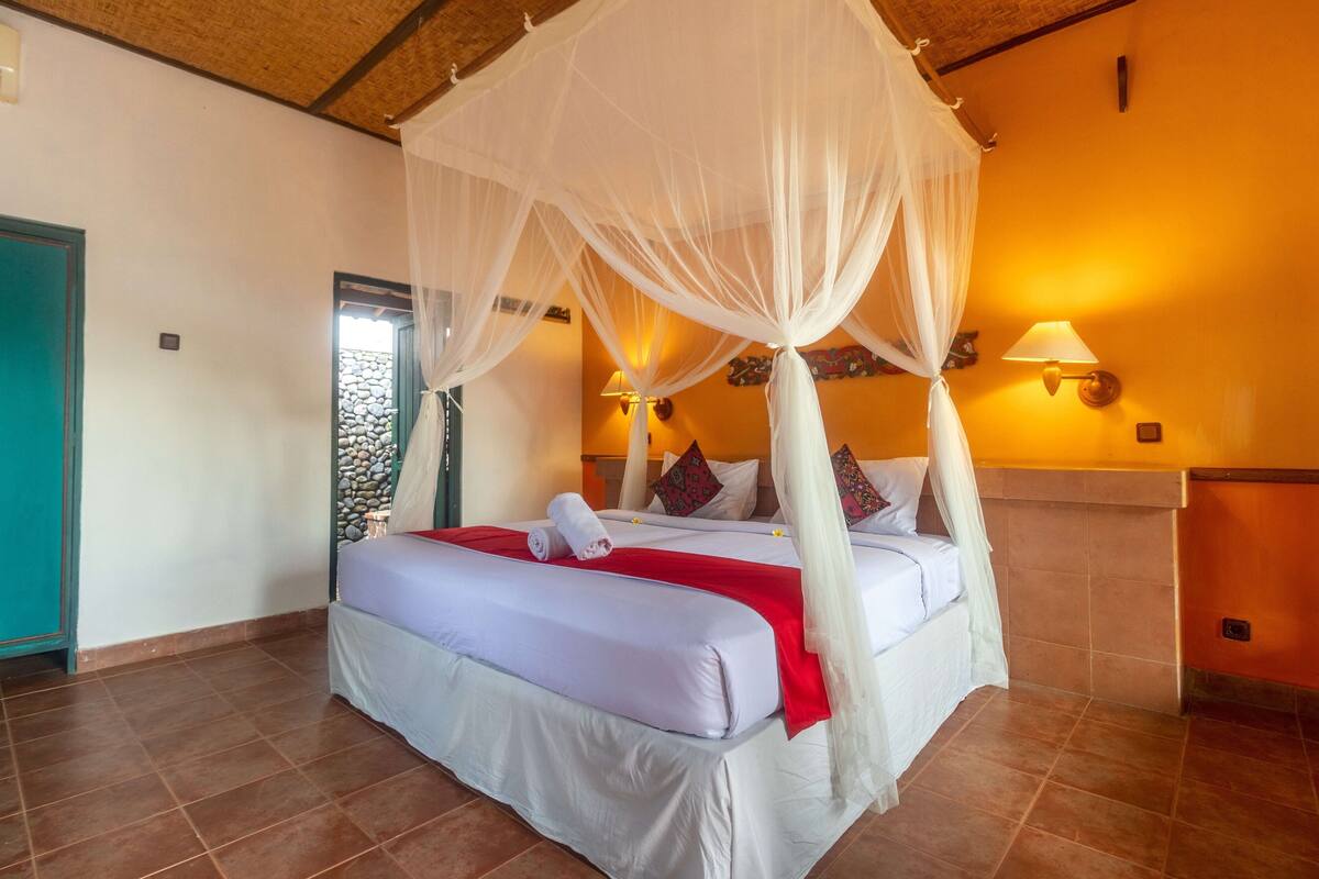 A bedroom with a four-poster bed, white linens, a red throw blanket, and a canopy.