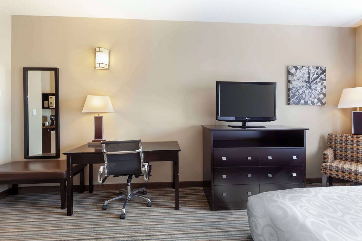 Ein Hotelzimmer mit einem Bett, einem Schreibtisch mit Stuhl, einem Fernseher auf einer Kommode, einer Lampe, einem Spiegel und einem Wandbild.