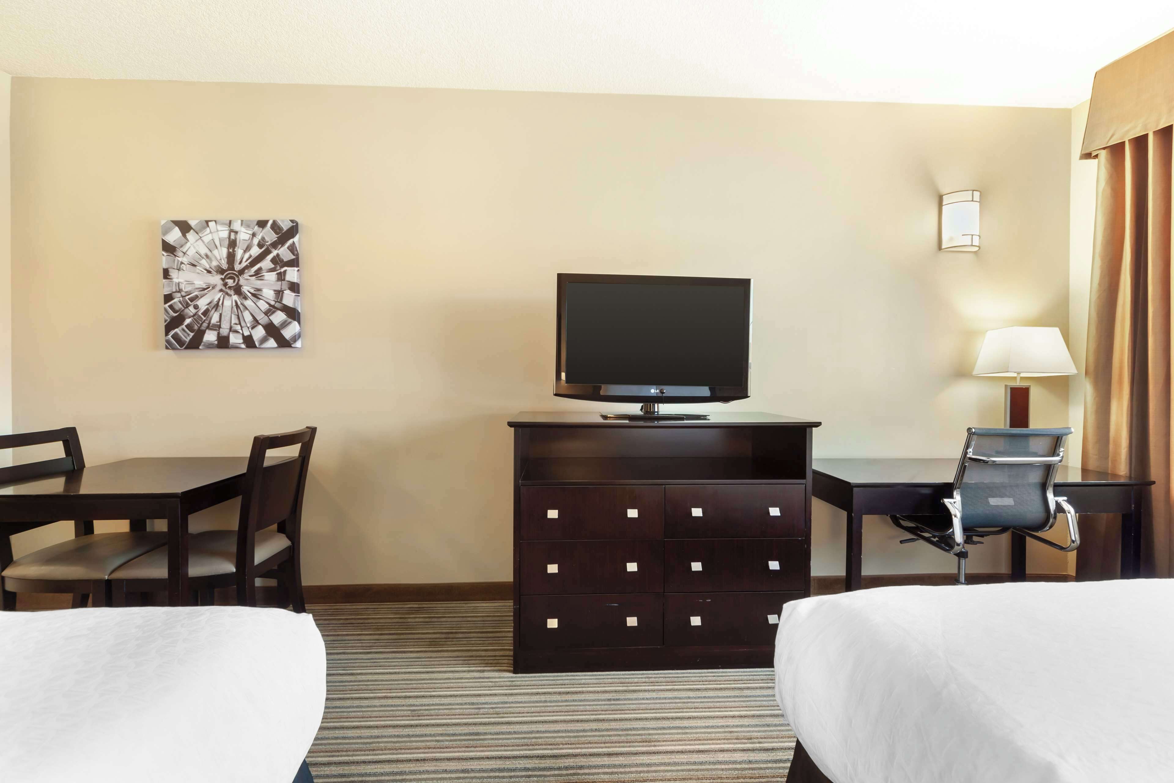 Habitación de hotel con dos camas, un escritorio con silla, televisión, cómoda, lámpara y una obra de arte en la pared.