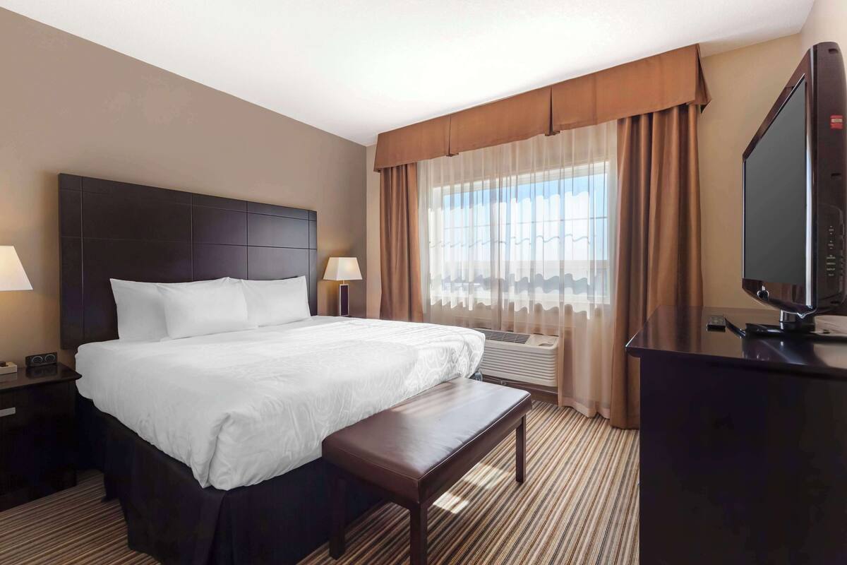 Ein Hotelzimmer mit einem großen Bett, einem Schreibtisch mit Fernseher, einer Bank und einem Fenster mit Vorhängen.