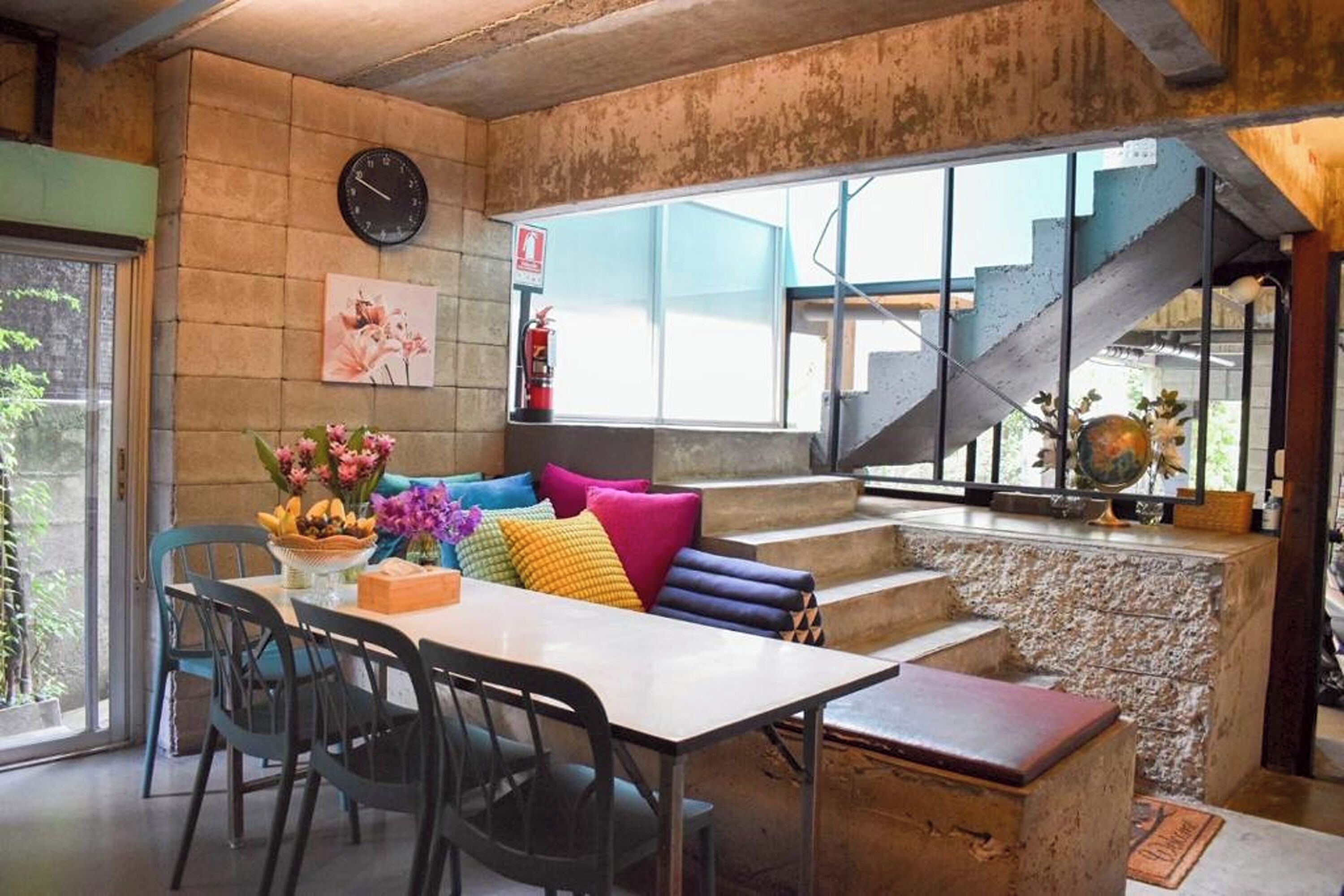 A dining area with a table, chairs, colorful cushions, a clock, and a staircase leading to an upper level.