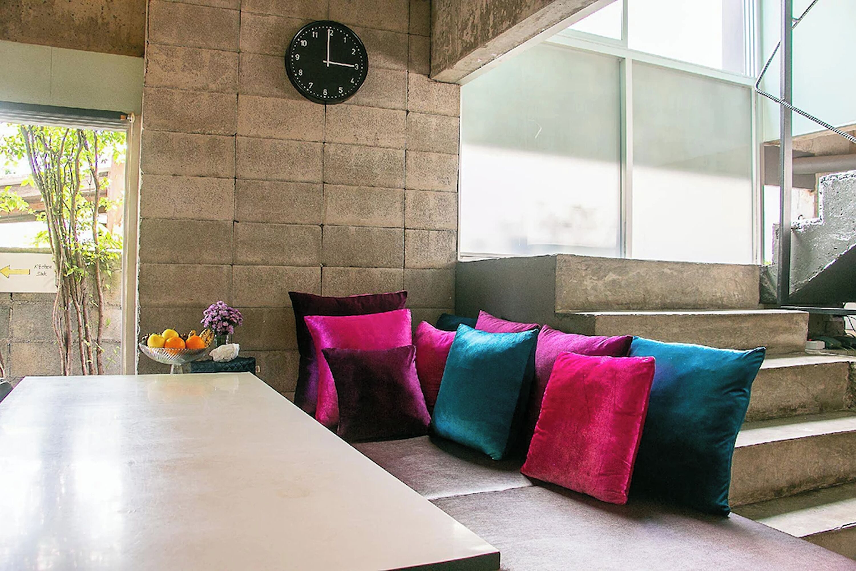 A modern interior with a large table, colorful cushions, a clock, and a window with a view of the outside.