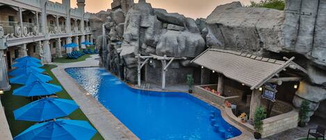 A swimming pool with blue umbrellas, a rock wall, and a building with balconies.
