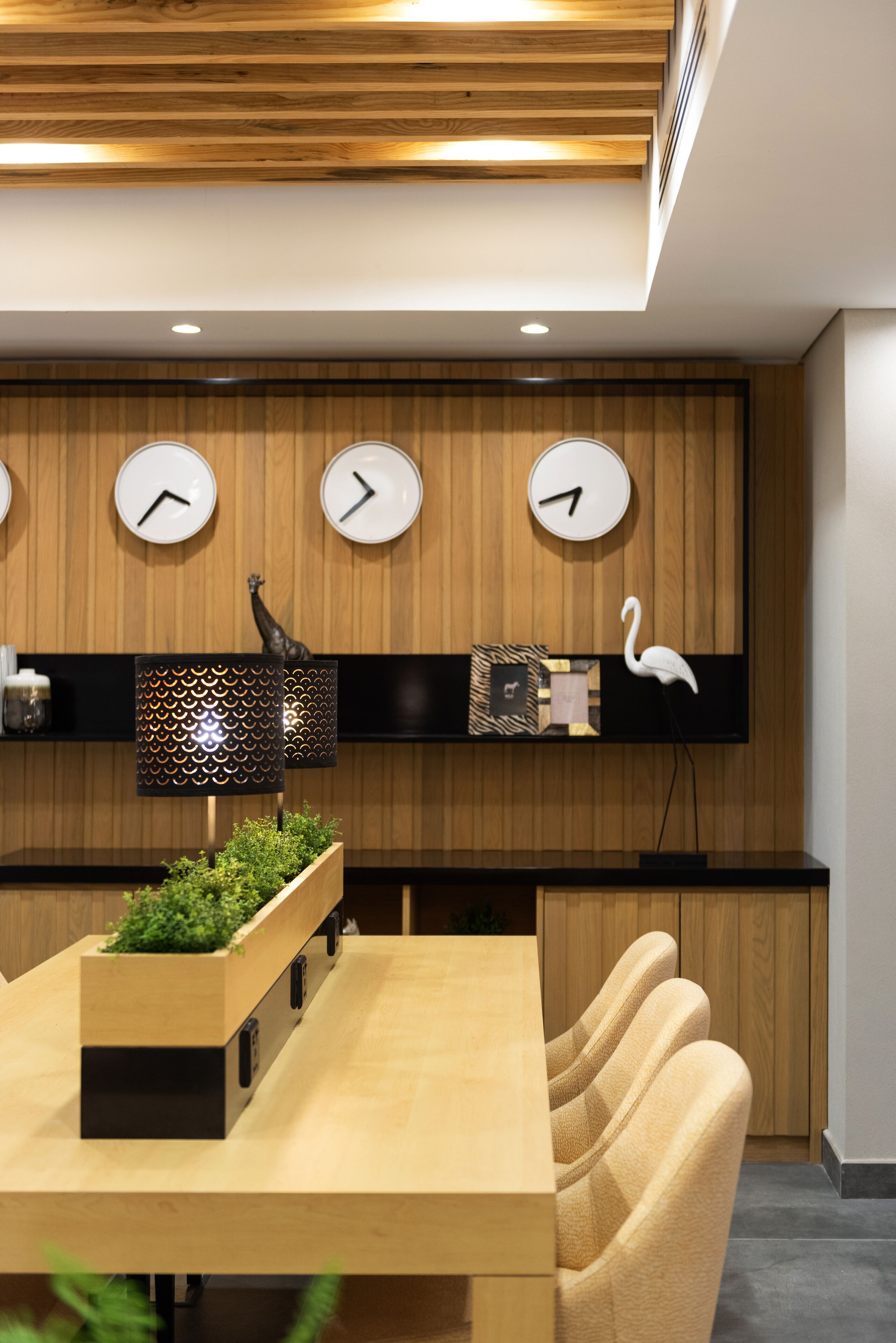 A modern kitchen with wooden panel walls, three wall clocks, a black shelf with decorative items, and a wooden dining table with chairs.