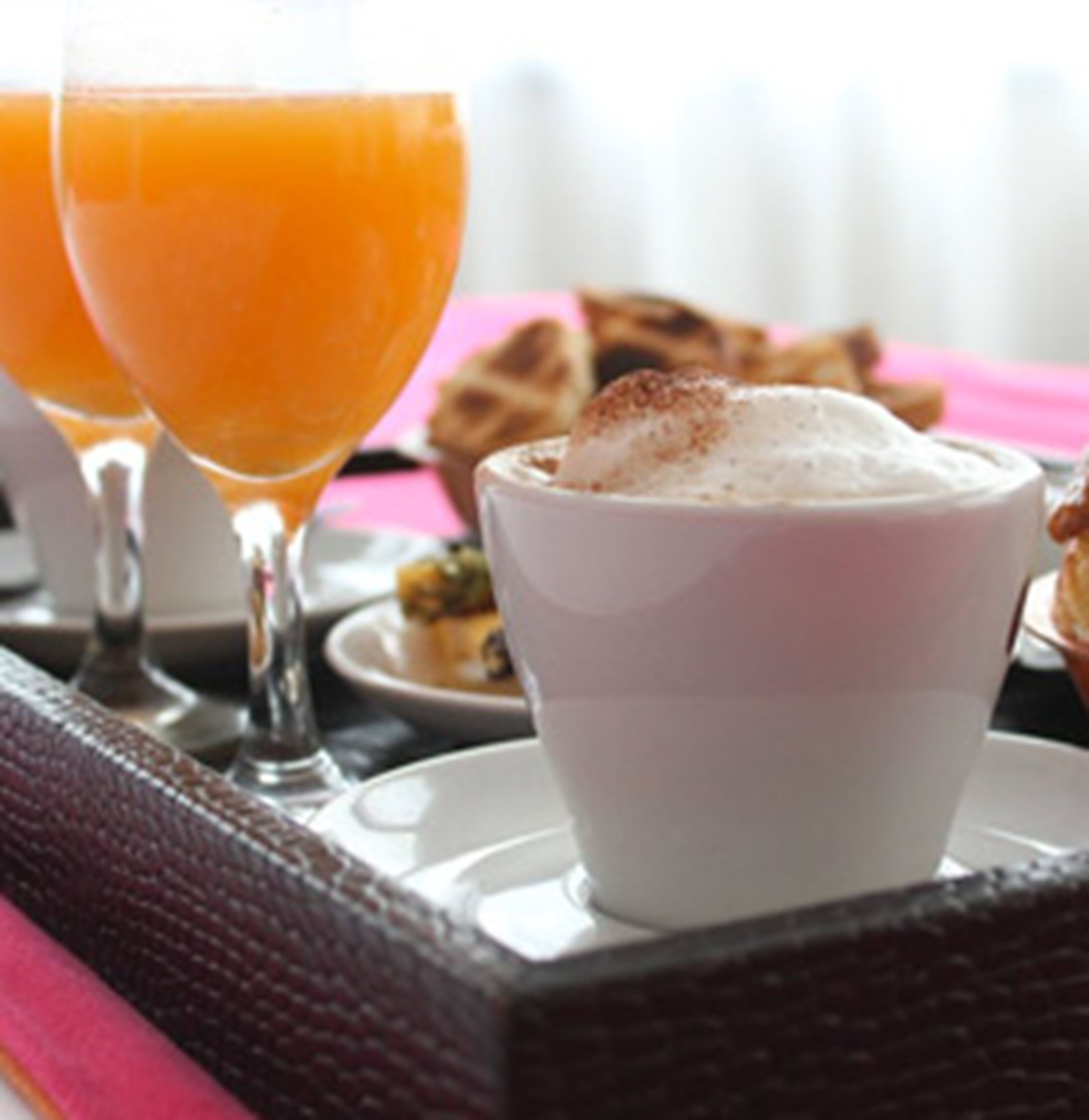 Une table mise pour le petit-déjeuner, avec un bol blanc contenant des aliments, deux verres de jus d’orange et une nappe rose.
