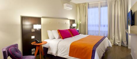 A hotel room with a bed, a chair, a small table, a TV, and a window with curtains.