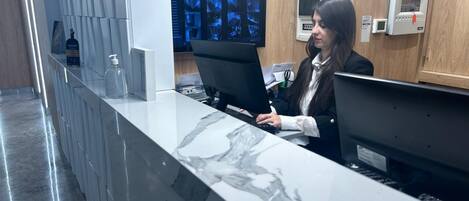 Balcão de recepção com tampo de mármore, dois monitores de computador e um painel de madeira ao fundo.