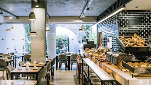 Un restaurante con una barra larga, varias mesas y sillas. Hay platos y cuencos en la barra, y se ve una parrilla al fondo.