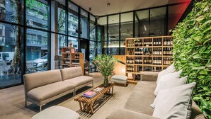 Un interior moderno con un sofá, un reposapiés y una mesa de centro de madera. Se observa una estantería llena de libros y un ventanal con vistas a la calle.