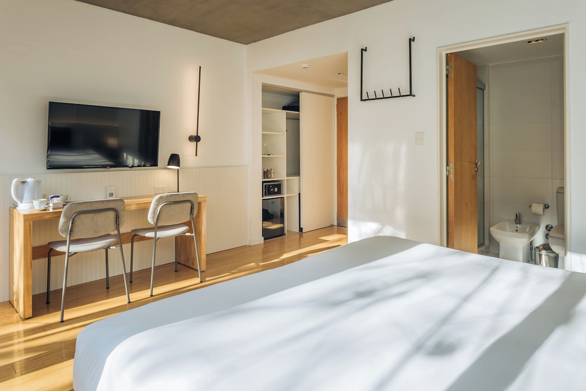 A modern hotel room with a large bed, a desk with two chairs, a TV, and a walk-in closet.