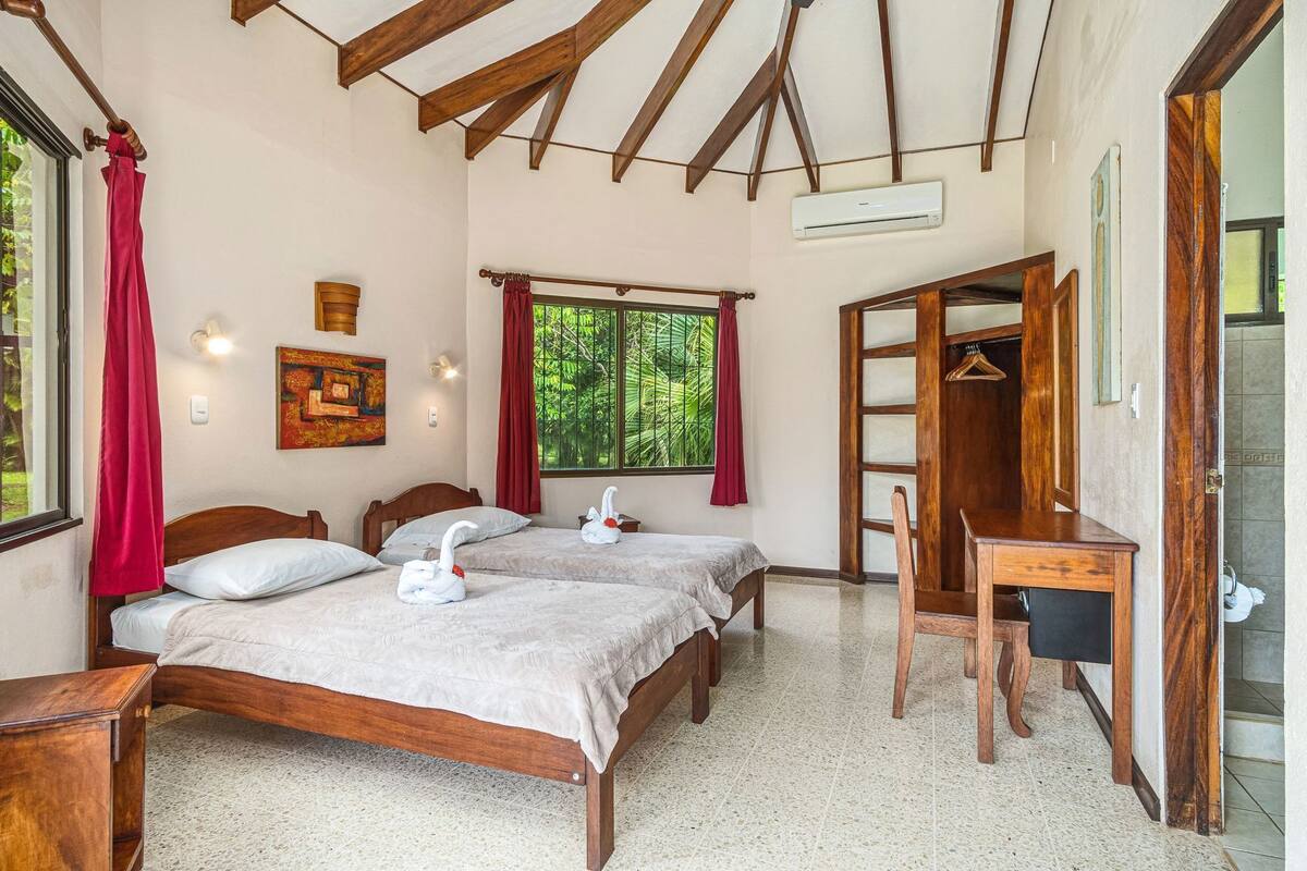 Una habitación con dos camas de madera, un escritorio, un armario y una ventana con vistas a la vegetación.