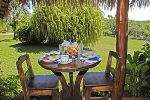A round wooden table with chairs, set outdoors with a lush green garden and a large palm tree in the background.
