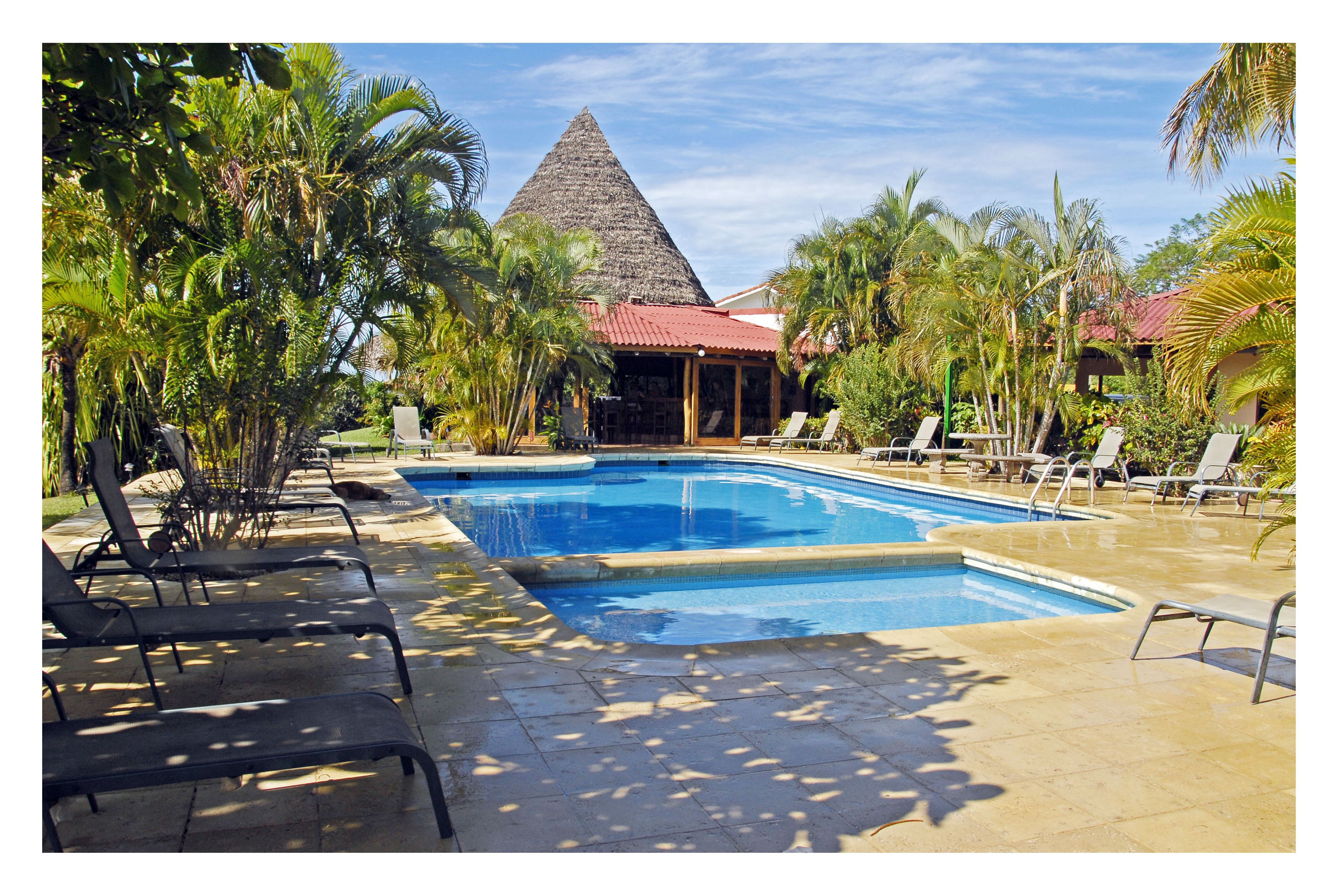 A swimming pool with lounge chairs, surrounded by palm trees and a building with a red roof.