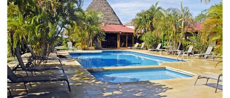 A swimming pool with lounge chairs, surrounded by palm trees and a building with a red roof.