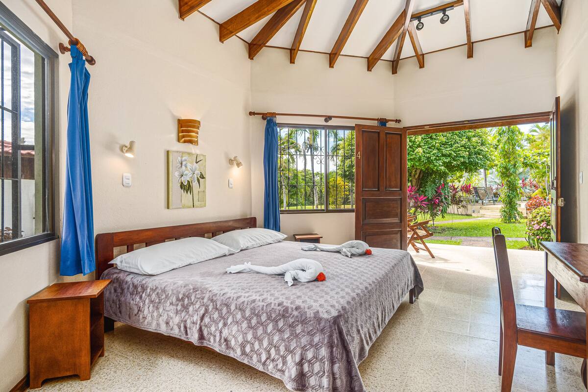 Un dormitorio con cama, cabecera de madera, mesitas de noche, un escritorio y vistas a un jardín a través de una puerta abierta.