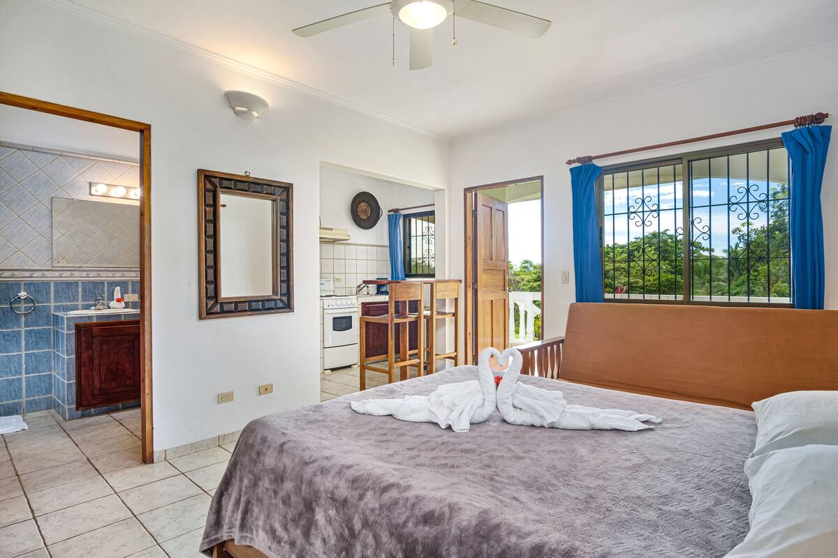 Una habitación de hotel con cama, baño, cocina y balcón con cortinas azules.