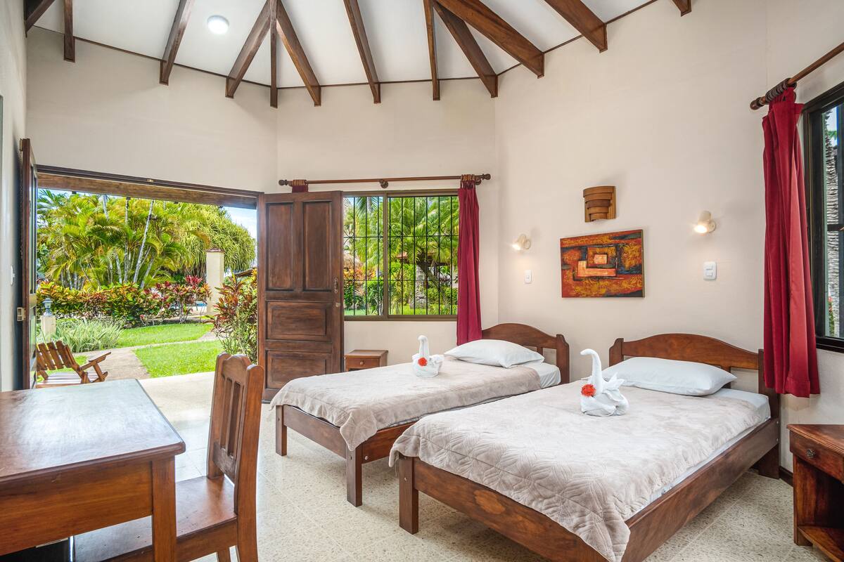 Habitación de hotel con dos camas de madera, una mesa de madera y vista a un jardín por un ventanal corredero.