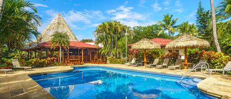 Una piscina con sillas de descanso y estructuras de techo de paja, rodeada de palmeras y plantas tropicales.