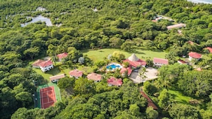 Luchtbeeld van een tropisch resort met meerdere gebouwen, een tennisbaan en een strand.