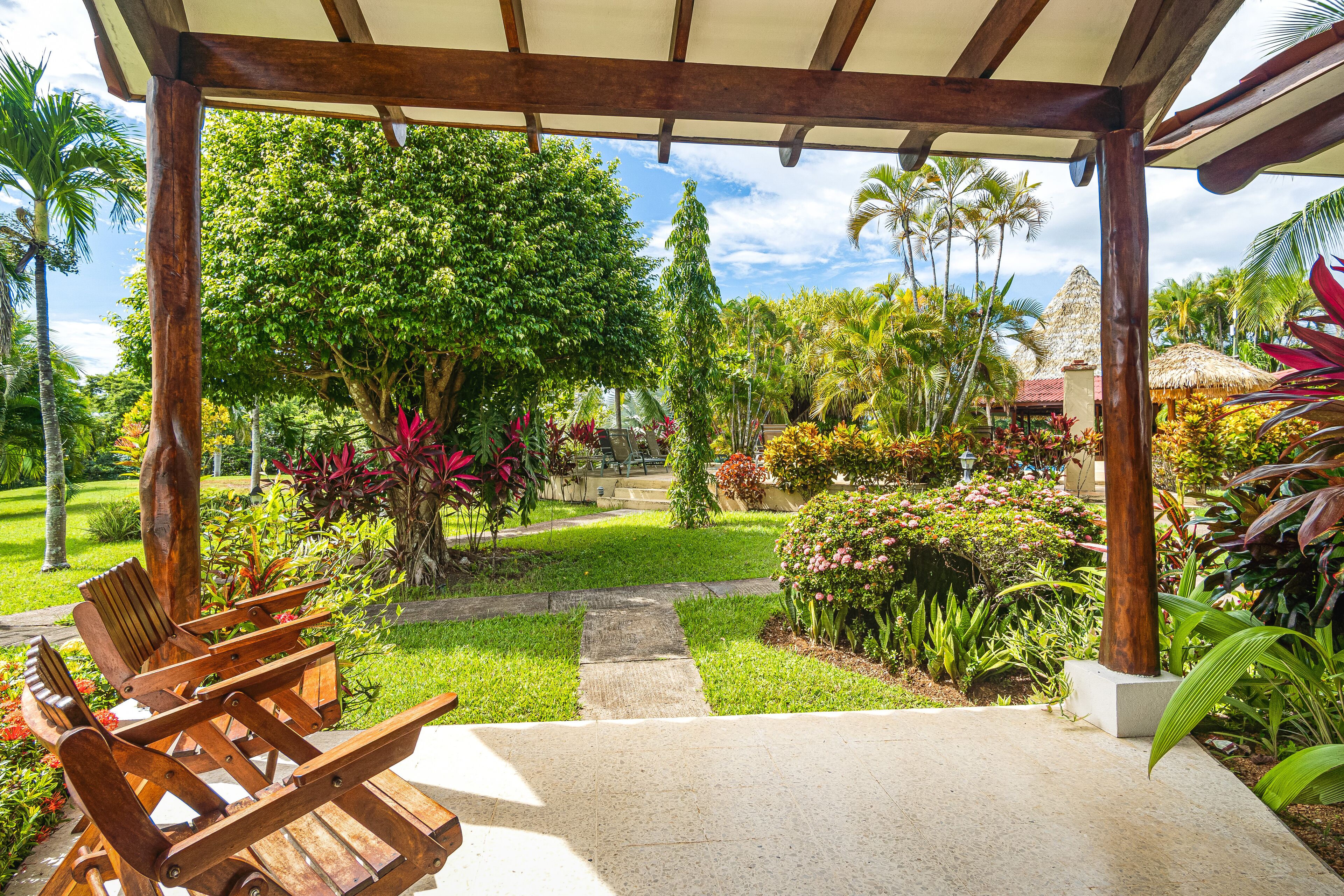 A covered outdoor seating area with wooden chairs, surrounded by lush greenery and a variety of plants.