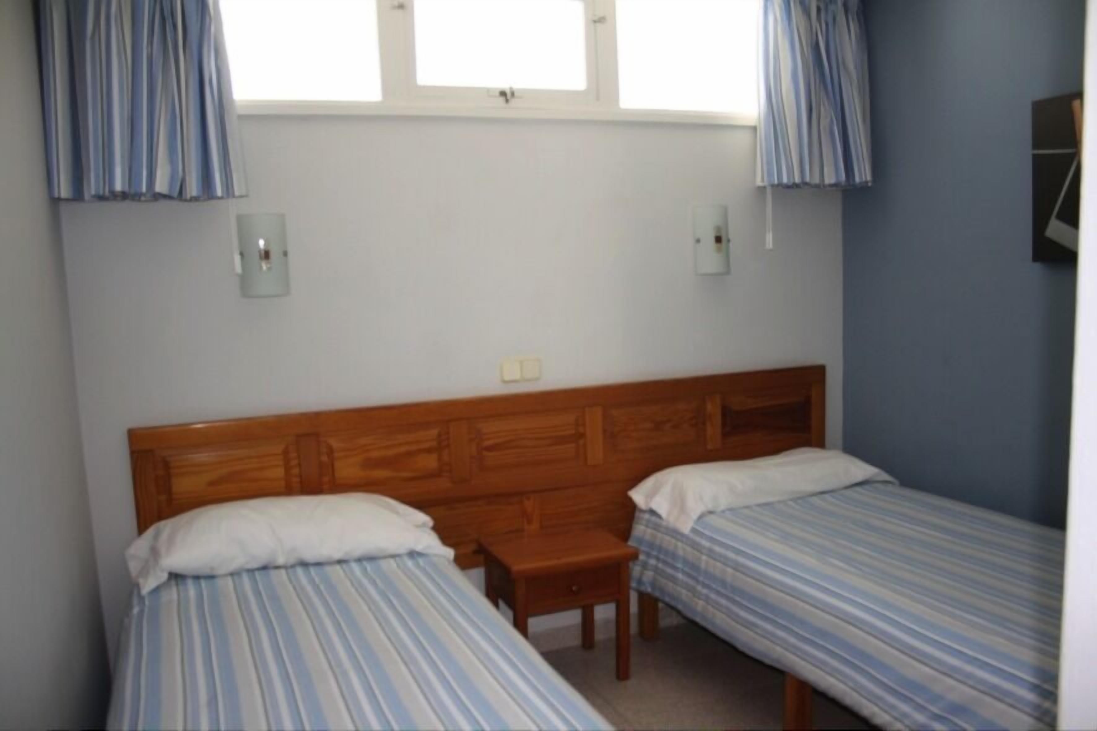 Een slaapkamer met twee eenpersoonsbedden, een houten hoofdbord, een klein nachtkastje en een raam met gordijnen.