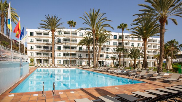 A swimming pool with lounge chairs and palm trees, surrounded by a multi-story hotel building.